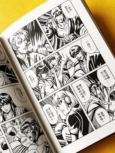Close-up of a physical comic book held open, showing detailed black and white inked panels, heavy paper texture, warm studio light.