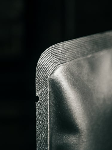 Angled studio close-up of a die-cut sachet edge being examined under manufacturing conditions, strobe lighting from the right illuminating the precise die-cut seam and foil surface texture, dark industrial background, focus on the mechanical sealing detail along the custom contour
