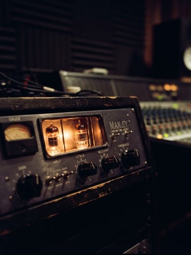 Moody close-up of analog tube gear glowing with warm amber light inside a dark, acoustically treated studio control room, shallow depth of field, cinematic 35mm film