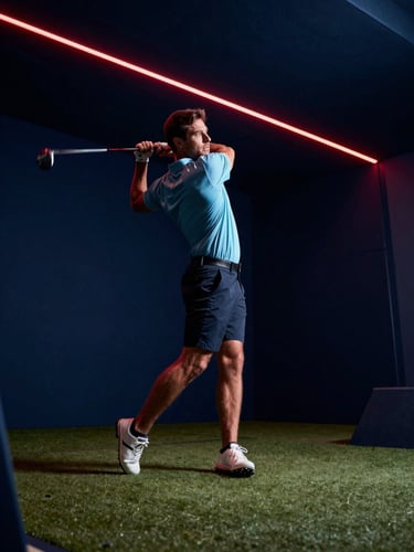 Low angle shot of a golfer mid-swing in an indoor simulator bay. Moody midnight blue and vibrant crimson LED overhead lighting. Dramatic motion blur on the golf club, high-contrast athletic atmosphere.