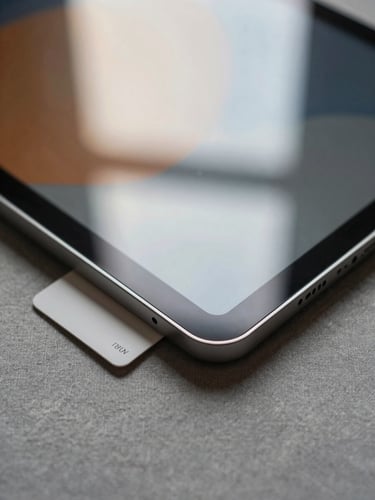 Extreme close-up overhead of a tablet edge and display corner resting on a warm grey linen surface, the display glass catching diffuse north-facing daylight, the aluminium frame edge sharp and precise, a Pantone swatch card half-tucked beneath the device corner — no UI visible on screen, soft warm ambient light, no flash