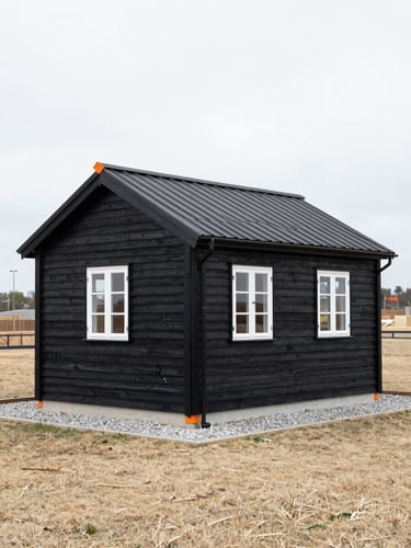 A classic black wooden summer house with white windows standing on a clean, dry lawn with a newly installed gravel drainage path along the foundation, overcast daylight.