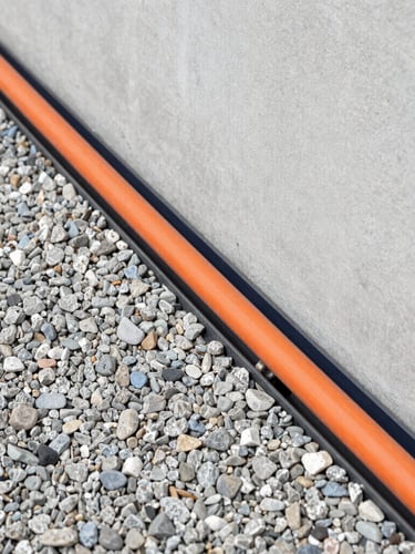 Close-up of a professional perimeter drain installation, orange drainage pipes laid in a clean gravel bed next to a residential concrete foundation, crisp overcast daylight, 35mm lens