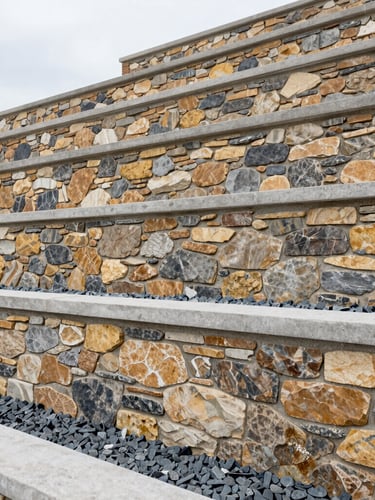 Close-up architectural shot of a multi-tiered stone and concrete retaining wall holding back a terraced slope, precise drainage gravel visible at the base, clean modern lines, overcast daylight, 24mm
