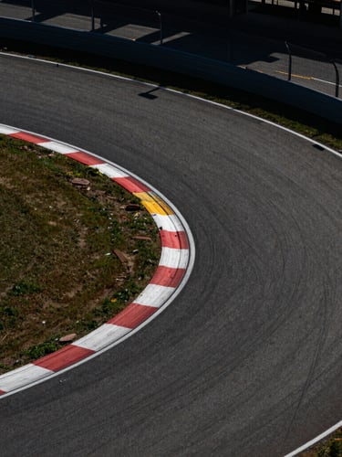 Dramatic top-down aerial shot of a racetrack's sharp asphalt curves, harsh midday sun casting sharp shadows, high-contrast industrial composition.