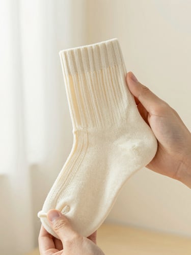 Close-up of hands holding a soft cream-colored woven sock, showcasing the detailed texture and precise stitching under soft side window light