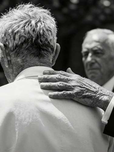 Fotografia in bianco e nero, un momento spontaneo durante un rito: le mani rugose di un anziano che accarezzano la spalla dello sposo, luce naturale di taglio, contrasto profondo.