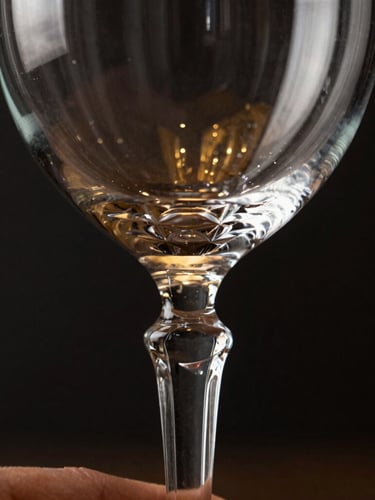 Macro close-up of a hand gently holding the stem of a crystal wine glass, showcasing the intricate detail and clarity of the glass, dramatic low-key studio lighting, sharp highlights on the glass edges, dark background, gold reflections, 85mm lens, focus on texture and craftsmanship.