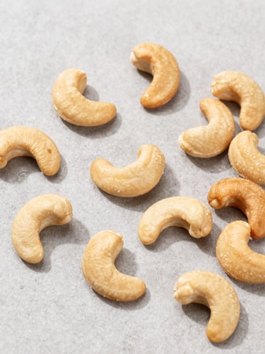 Overhead flat-lay of sorted premium whole cashew nuts on a clean, light-grey stone surface, bright natural daylight, sharp focus