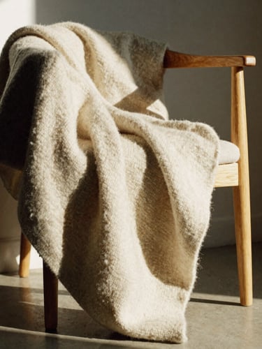 A heavy raw wool blanket draped over a minimalist oak chair, warm afternoon sunlight casting deep textures and high-contrast shadows, 35mm photography