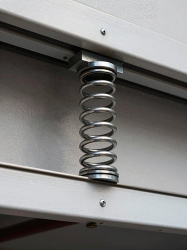 Straight-on architectural detail of a heavy-duty steel torsion spring mounted above a garage door frame, crisp daylight highlighting the metallic texture and precise coils, clean industrial aesthetic.