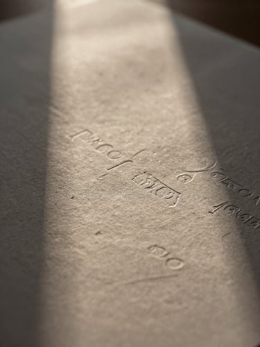 Macro photograph of heavy textured paper with deep letterpress indentation, soft directional window light casting long shadows across the tactile surface