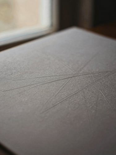Macro close-up of heavy textured archival paper with blind-debossed geometric lines, soft directional window light, muted bronze tones, shallow depth of field, 35mm film