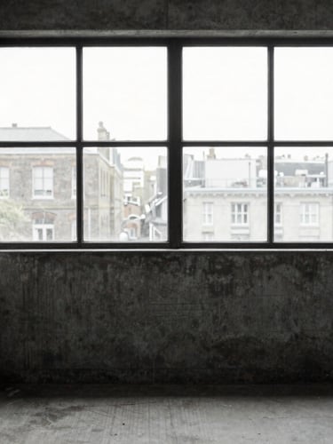 A quiet, high-contrast portrait of an empty concrete workspace in East London, overcast daylight pouring through a large steel-framed window, desaturated desaturated tones, 35mm film.