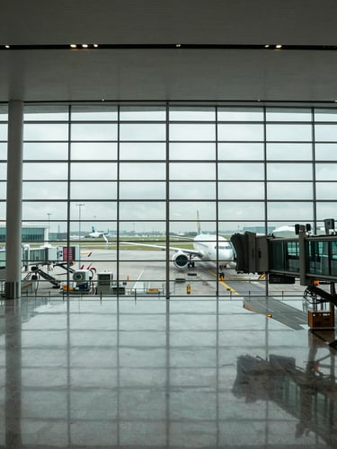 Wide panoramic view looking down a modern airport terminal concourse, gates receding into the distance on both sides, cool overcast daylight flooding through floor-to-ceiling windows, a single aircraft visible at a jet bridge to the right third of the frame, expansive polished floor reflecting the structural ceiling grid, no people