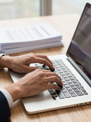 A close-up shot of professional hands typing on a modern laptop keyboard in a sunlit office environment in Côte d'Ivoire. Crisp physical documents sit neatly stacked on the side, shot with a 35mm lens, warm and welcoming lighting.