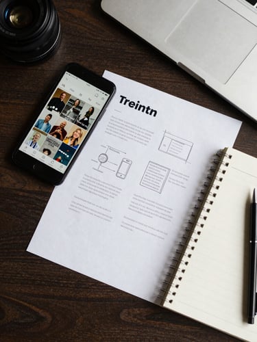Overhead flat-lay of a desk with a phone showing social media graphics, a printed brand sheet, and a notepad with sketched layout ideas, clean daylight from above