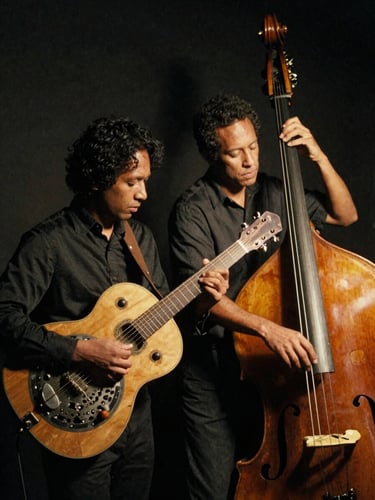 Low-key atmospheric portrait of the blues duo with resonator guitar and upright bass, warm side spotlight, deep charcoal shadows, 35mm film grain.