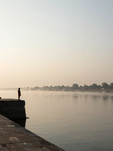 Wide panoramic view of the Ganga at early dawn, mist rising off still water, a lone silhouette of a pilgrim standing at the ghat's stone edge to the left third, vast pale sky above, diffused golden light on the river surface
