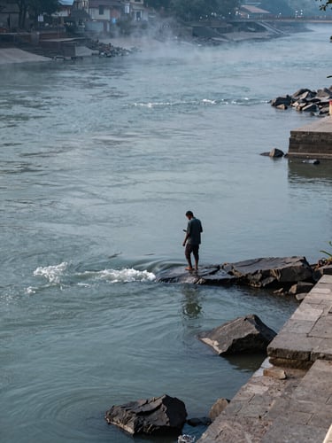 Wide documentary frame, a solitary pilgrim standing waist-deep at the rocky Devprayag confluence where two rivers meet, early morning diffused light, mist rising from the water, stone ghats visible at the far right edge, cool blue-gray tones