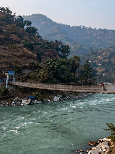 Wide shot of Rishikesh Laxman Jhula suspension bridge at dawn, Ganga flowing below in pale blue-green, forest-covered hills rising steeply behind, one lone figure crossing the bridge, soft early light