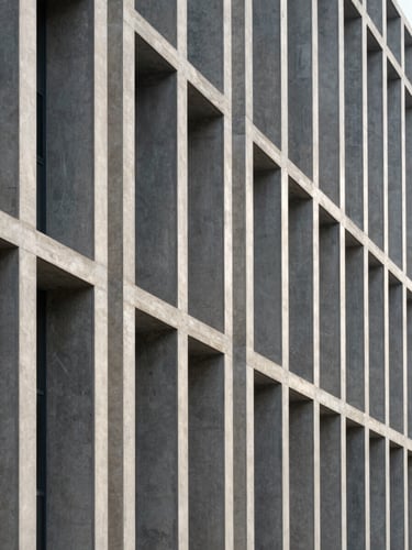 A high-contrast, wide architectural shot of a modern concrete and steel building facade. Sharp diagonal shadows cut across clean vertical lines. Cool morning natural light emphasizes the structural grid.