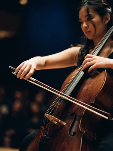 Intimate close-up of a young cellist's hands positioning a bow on strings, warm concert hall stage lighting, deep midnight blue background, shallow depth of field, 35mm