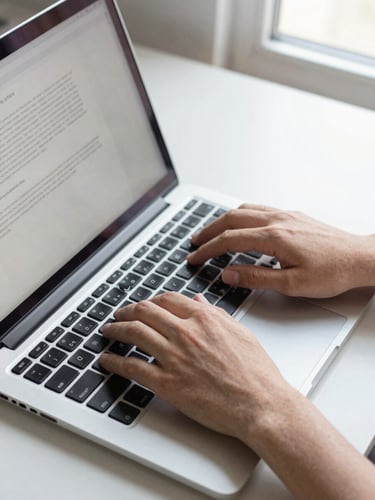 Overhead close-up of hands typing on a laptop keyboard with a brief document visible on screen, clean desk surface, daylight from a window to one side, no people's faces