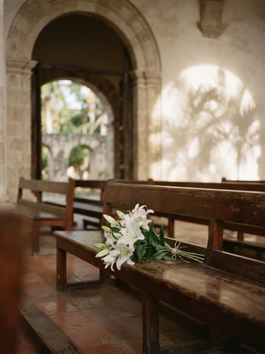 Soft afternoon light filtering through the high arch of a serene chapel, delicate white lilies resting on a polished wooden bench, palm shadows on a warm off-white wall, Dominican Republic.