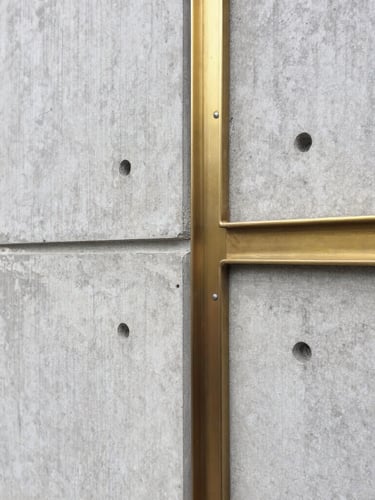 Sober architectural photography of a raw concrete wall meeting a brushed brass structural joint, natural diffused daylight, sharp focus