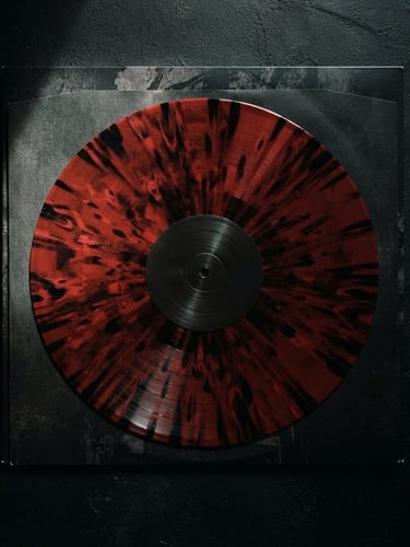 Top-down flat lay of a splatter vinyl record, dark blood-red and black wax, resting on a heavy textured gatefold jacket, dramatic chiaroscuro lighting, harsh shadows, 35mm film.