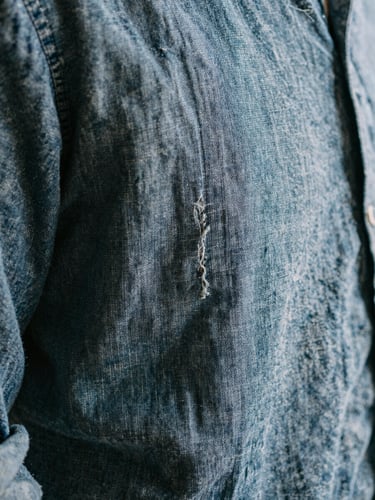 Macro detail of a well-worn, washed indigo linen shirt seam, showing soft frayed edges and textured weave under diffused daylight.