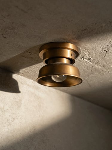 Macro shot of a heavy sand-cast bronze downlight installed in a raw concrete ceiling, harsh direct sunlight casting a sharp geometric shadow across the plaster wall.