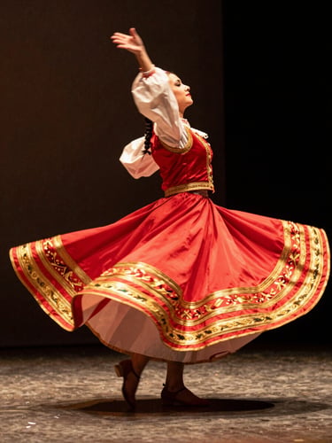 Low-angle dynamic shot of a folkloric dancer in mid-spin, vibrant crimson and gold embroidered skirt catching warm natural studio light, subtle motion blur, 35mm film grain.