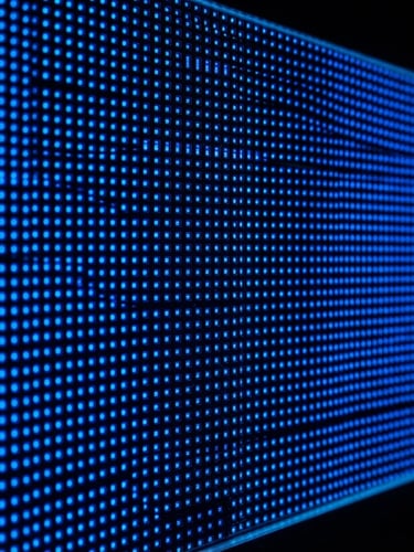 Close-up of a commercial LED display panel showing vibrant electric blue pixels, set against a dark background with a subtle edge-light, sharp focus on the panel's surface, wide aspect ratio