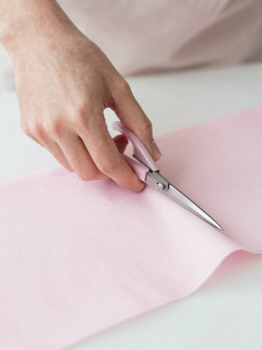 Close-up of hands carefully cutting light pink fabric with tailor's scissors on a white cutting mat, soft diffused morning light, delicate pastel tones, shallow depth of field, 50mm, soft editorial photography