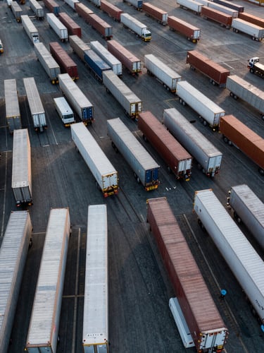 Aerial drone perspective of a busy European cargo dock at dawn, rows of refrigerated and dry freight trailers staged in parallel bays, overhead overcast light casting flat even illumination, no workers visible, infrastructure and vehicle scale dominating the frame