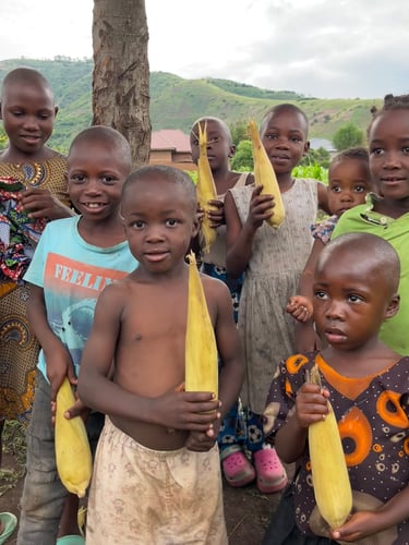 A group of smiling African children holding fresh ears of corn in a rural village setting.