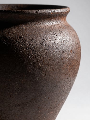 A high-contrast profile of a coarse, iron-speckled ceramic vessel with heavy kiln scars, cast in deep shadow with dramatic side-lighting from the right.