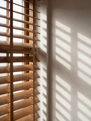 A close-up of warm sunlight filtering through half-open wood Venetian slats, creating clean graphic shadow lines on a minimalist plaster wall, 35mm lens.