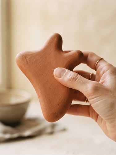 Macro detail shot of a matte terracotta sculptural wellness device, held gently by a hand, soft diffused natural window light, warm alabaster background.