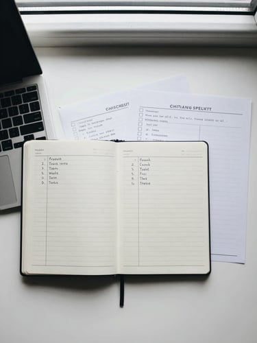 Overhead flat-lay of a clean desk: open planner notebook with handwritten task columns, a laptop keyboard edge entering from the left, two printed checklist sheets side by side, cool north-facing window light, minimal objects, no clutter