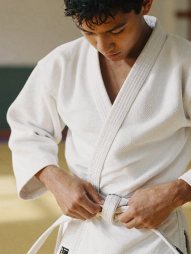 Dynamic candid close-up of a young athlete tying their white belt in a sunny dojo, focused expression, authentic documentary photography, natural daylight, warm earthy tones, 35mm film aesthetic.