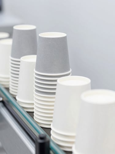 Crisp industrial product photography of neatly stacked white and gray paper cups on an automated conveyor line, bright high-key lighting, sharp focus.