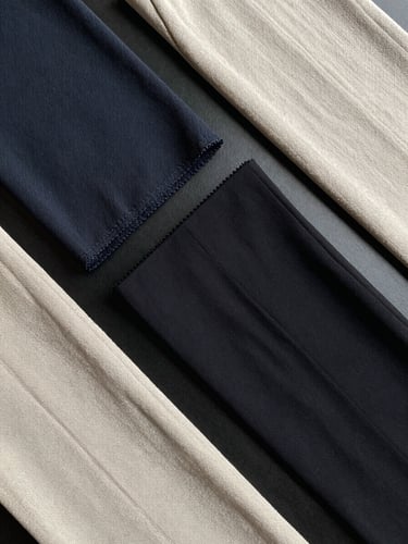 Overhead flat-lay arrangement of formal trouser fabric swatches — premium Barbie fabric, imported Lycra, and stretch linen — spread across a matte black surface, each swatch showing distinct weave texture under bright even studio lighting, no human subjects, no styling