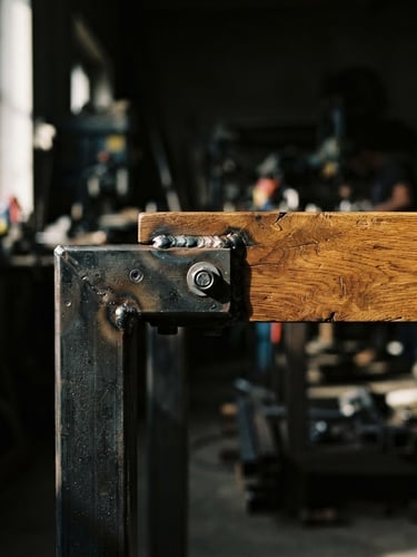 Gros plan sur un assemblage de table industrielle en bois de chêne massif et acier brut, avec des soudures apparentes et la texture du bois mise en valeur, sous une lumière latérale dramatique d'atelier, tons chauds et froids contrastés.