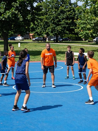 A volunteer coach in an orange athletic shirt instructing a diverse group of children on an outdoor basketball court, bright daylight, authentic Ohio park setting.