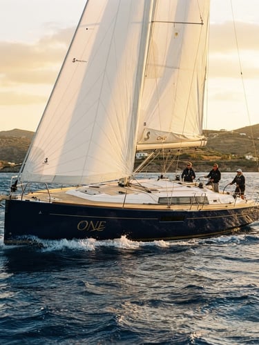 Wide environmental shot of the deep blue hull of the Oceanis 60 sailing yacht ONE slicing through deep blue Mediterranean water, bright golden-hour sunlight, white sails filled with wind, 35mm film look.
