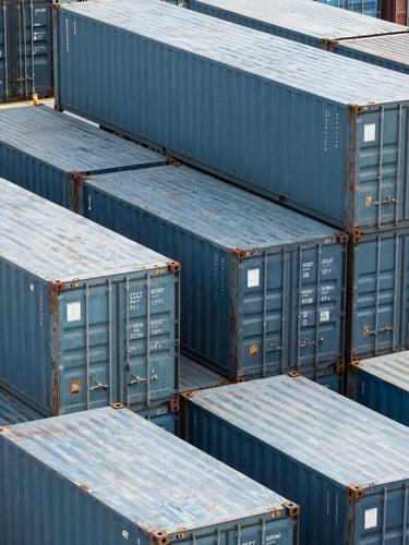 High-angle structured pattern of steel shipping containers stacked in a modern deep-water port, cool daylight with subtle blue accents, crisp commercial photography