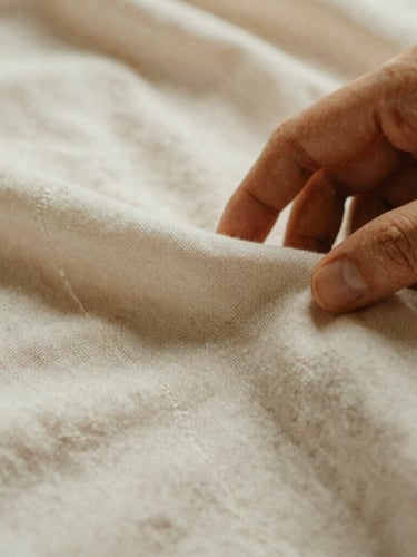 Close-up macro shot of a hand gently touching a breathable cotton fabric, showing the weave and stitching details, under warm, diffused natural daylight. Soft shadows, muted earth tones, 35mm film aesthetic.